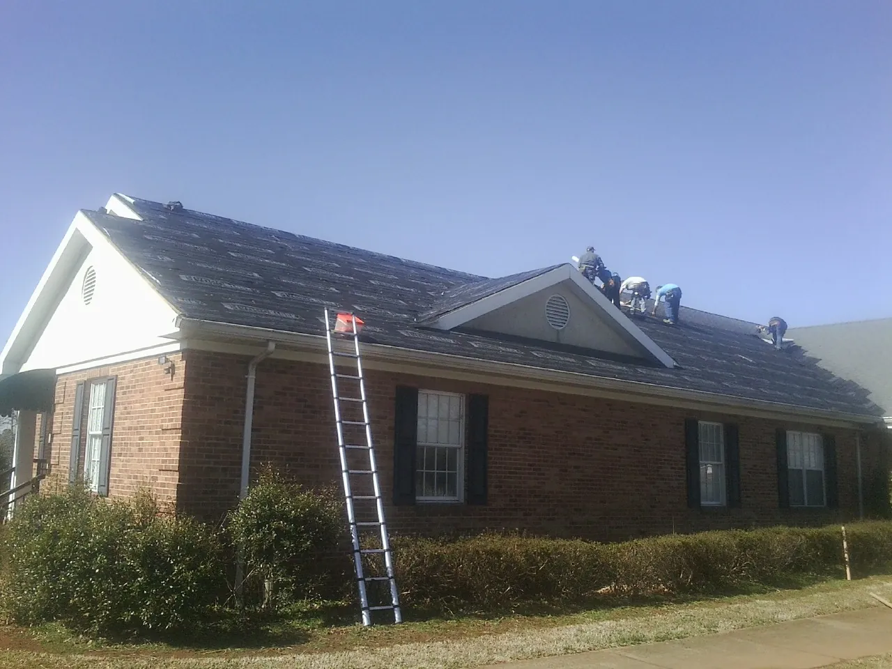 Crew tearing shingles off a brick ranch roof with a ladder set — Emergency Roof Repair work by our crew in Ulysses