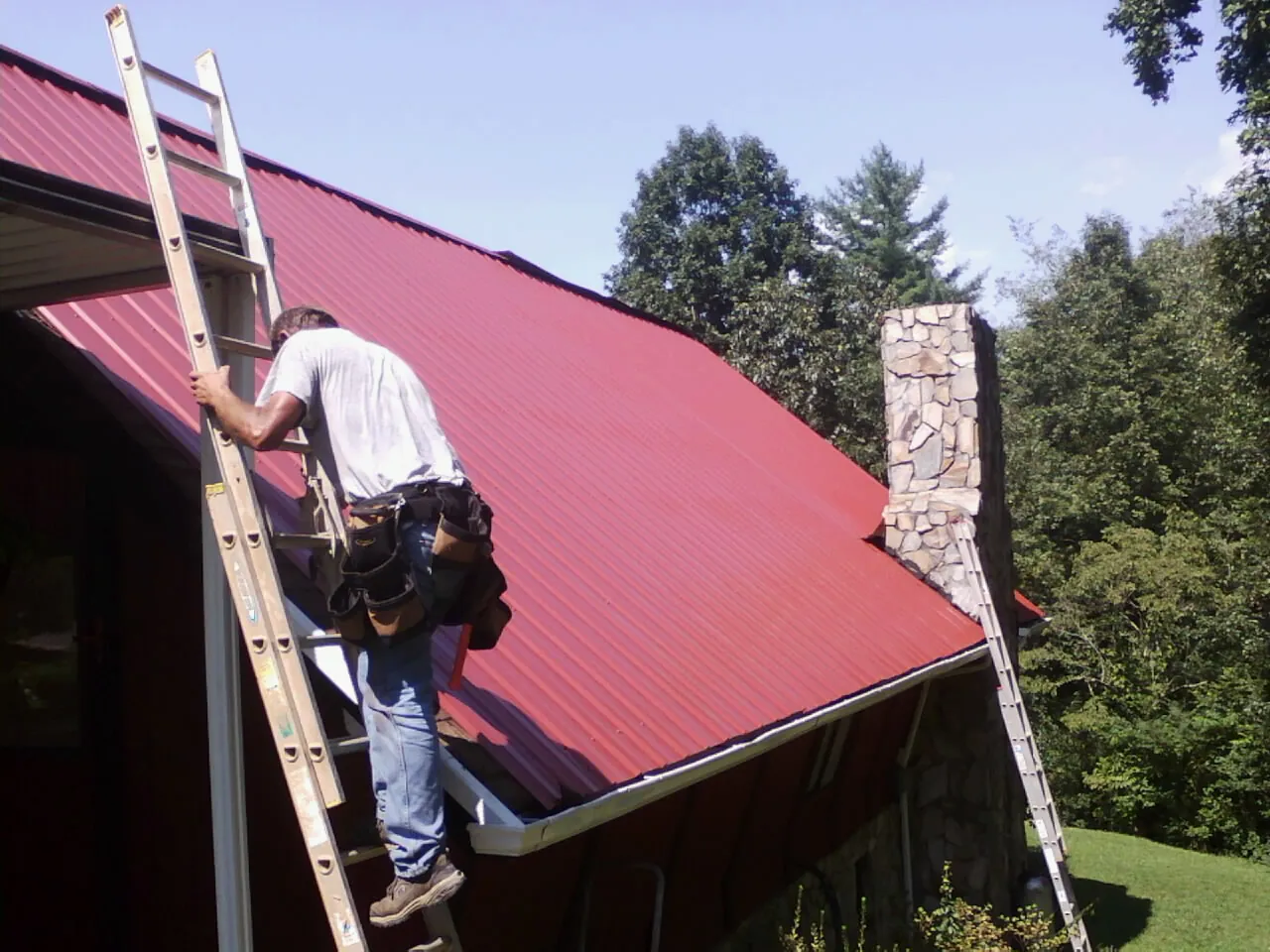 Roofer on a ladder beside a red metal roof and stone chimney — Soffit & Fascia Repair by Roofing Brothers in Ulysses